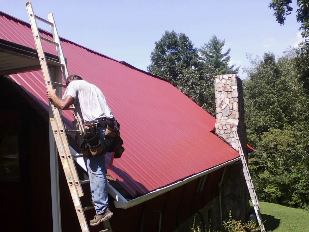 Roofer on a ladder beside a red metal roof and stone chimney — Soffit & Fascia Repair by Roofing Brothers in Sadieville